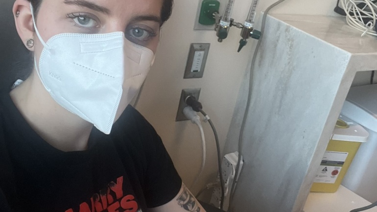 This picture is a selfie that Kate Hilts snapped when receiving a life-saving infusion as part of her treatment plan in the chemo ward at Sibley Memorial Hospital in Washington, D.C., on February 20, 2024. Kate, a white woman with dark brown hair and blue eyes wearing a white respirator and black Harry Styles t-shirt, is sitting on a hospital bed wiht an IV in her left forearm and a green bandage wrapping near her forearm. medical machines, cords, and the plugs they go into on the hospital wall can be see in the background. The IV is dispensing a life-saving medication. If paid out of pocket, the medication alone would cost nearly $100,000.