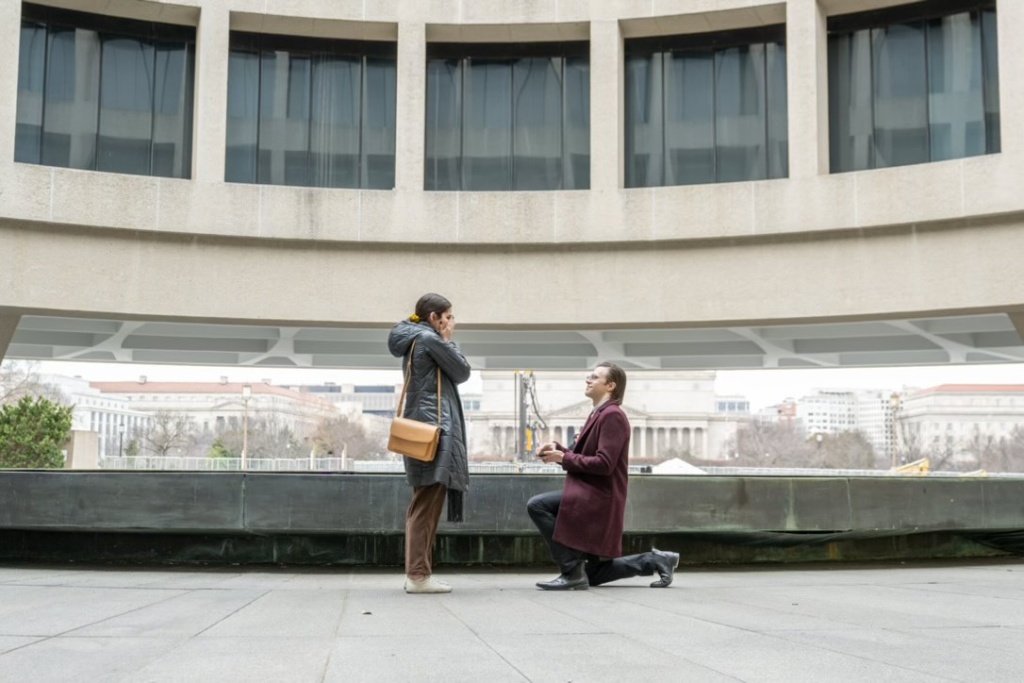 A picture of Kate’s longtime partner, Ross, proposing to Kate outside the renowned Hirshhorn Museum and Sculpture Garden in Washington, D.C., on February 24, 2024. They are in front of a fountain. Ross, Kate's longtime partner, is on the right side of the picture on one knee, holding open a ring box to Kate. Ross, a white man with glasses and brown hair brushed back and touching the top of his shoulders, is looking up at Kate with an expression of joy and love. Ross is wearing a plum colored coat, black slack, and black boots. Kate, a white woman with her dark brown hair pulled back with a yellow scrunchy, is on the left side of the picture in brown slacks, white tennis shoes, a shiny grey winter coat with a tan-colored rectangular bag. Her hands are covering her face and she is conveying a clear reaction of delight surprise.